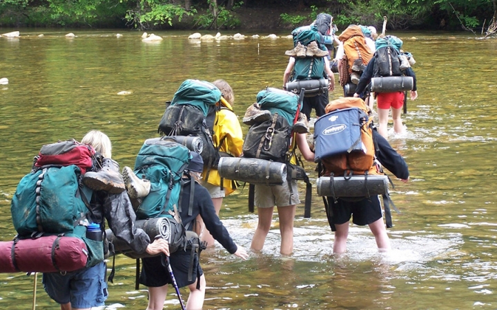 A group of people wearing backpacks cross a river in ankle-deep water. 